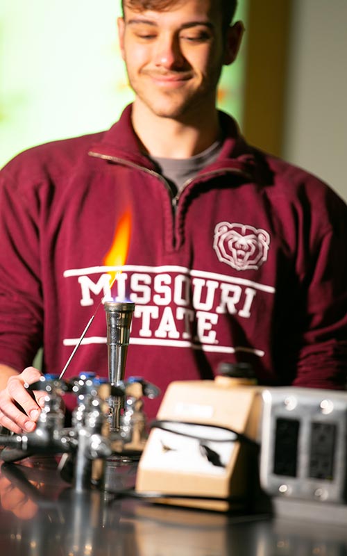 A student in a Missouri State hoodie stands behind a lit Bunsen burner.