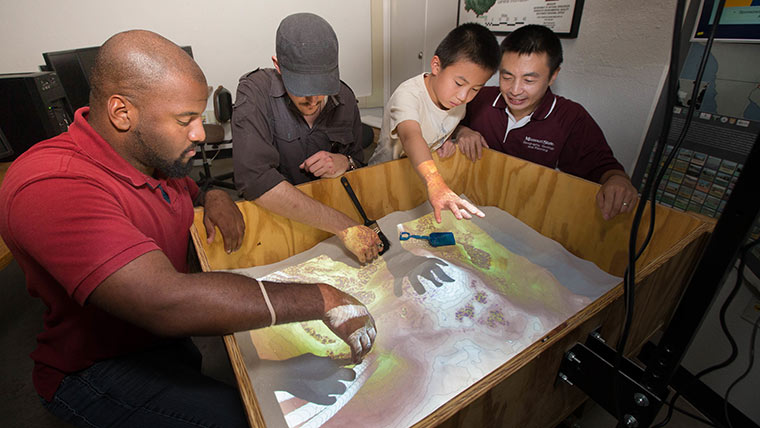 A professor, two graduate students and the professor's young son use an augmented reality (AR) sandbox.