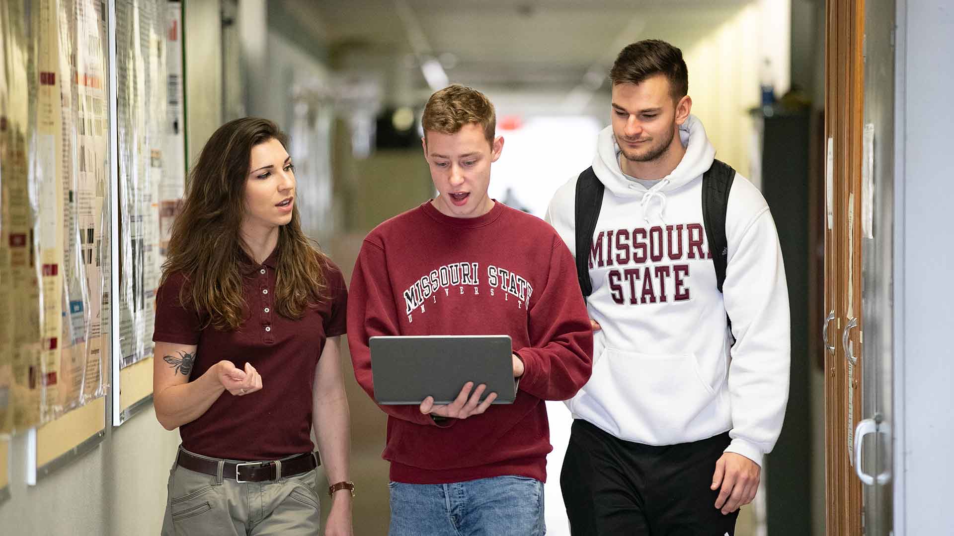 Three students in BearWear looking at a laptop as they walk down a hallway in Temple Hall.